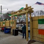 Wooden pergola structure with green flags, American flag flying overhead on a cloudy day.