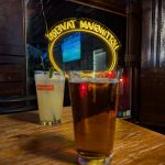 Two drinks sit on a wooden bar counter in front of a neon sign reading "MANHATTAN."