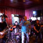 Band performing live on a rustic stage with musicians playing instruments.