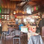 Cozy, rustic bar interior with wooden ceiling, stocked shelves, and patrons enjoying drinks.