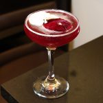 Red cocktail in a stemmed glass, garnished with white and red edible flowers on a dark surface.