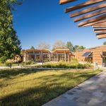 Modern wooden buildings with glass windows set in a sunny, grassy landscape under a blue sky.