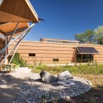Modern wooden building with glass facade and natural landscaping under a clear blue sky.
