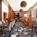 Modern restaurant interior with warm wood tones, high ceilings, and large windows.