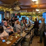 Busy pub interior with bar, patrons socializing, and warm overhead lighting.