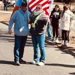 Two men holding American flags walk down a sunny, paved road.