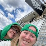 Two women in green hoodies smiling outdoors with a "Tir Na Nog" sign overhead.