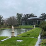 Formal garden with a glass pavilion, reflecting pool, and manicured lawns under an overcast sky.