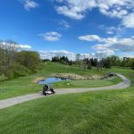Golf cart on a green course with pond and rolling hills under blue sky.