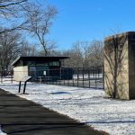 Winter park scene with a modern pavilion, snow-covered ground, and bare trees under a blue sky.