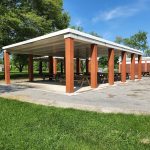 Open-air pavilion with concrete roof and brick pillars, set on a grassy area under blue sky.