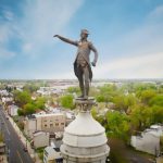 Statue of a man pointing atop a white pedestal, set in a lush, green city park under a blue sky.