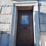 Bronze door with barred window and historical plaque, set in a stone building entrance.