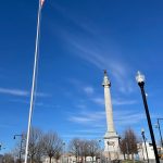 American flag flies high above a tall monument under a bright blue sky.