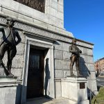 Bronze statues guard the entrance of a classical stone building under a clear blue sky.