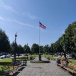 American flag flies over a paved walkway lined with benches and trees on a sunny day.