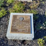 Bronze memorial plaque set on a granite base in a landscaped outdoor area.