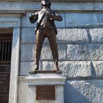 Bronze statue of a man in period dress stands before a large stone monument.
