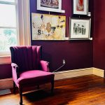Deep purple wall gallery with framed art, featuring a pink velvet armchair on dark wood floor.