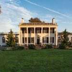 Elegant white neoclassical manor house with columns and manicured lawn under a blue sky.