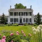 Neoclassical white manor house with columns, set against a lush green lawn and blooming garden.