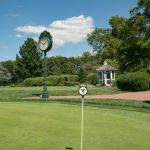 Golf course scene with a clock post, flagstick on the green, and clubhouse in the background.