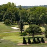 Golf course green with manicured fairways, trees, and distant wooded hills under a bright sky.