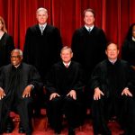 Group of diverse individuals in black robes posing on a stage with red curtains.