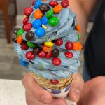 Colorful, swirled blue ice cream cone topped with candy and sprinkles held in hands.