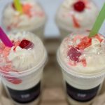 Three cups of creamy, colorful dessert topped with whipped cream and sprinkles on a wooden tray.