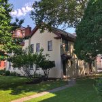 Historic light-colored house nestled among large, mature green trees on a sunny day.