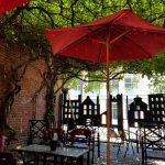 Red umbrella under green trees over outdoor cafe seating against a brick wall.