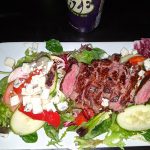 Sliced steak served on a bed of mixed greens with tomatoes, cucumbers, and feta cheese.