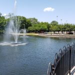 Fountain spraying water in a large pond surrounded by trees and a park walkway.