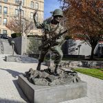 Bronze statue of a soldier in uniform and helmet standing on a rocky base outdoors.