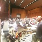 Orchestra rehearsal in a large hall with musicians and brass instruments visible.