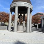 Bronze statue under a large, domed monument structure on a paved plaza.