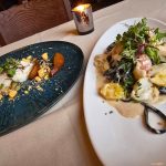 Gourmet dish with seafood and greens on a white plate, next to appetizers on a dark blue platter.