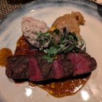 Sliced steak with a rich sauce, topped with a grain salad and fresh herbs on a white plate.