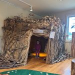 Child stands inside a large, rustic fort made of branches and wood indoors.