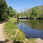 Scenic view of a calm river flowing under a wooden bridge, lined by trees and a dirt path.