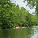 Kayakers paddle down a calm, tree-lined river under an overcast sky.