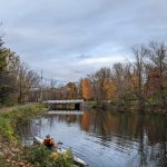 Kayaker on a small inflatable boat on a calm river beneath an overcast sky with trees in autumn colors.