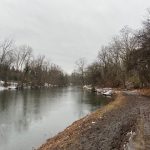 Winter landscape of a calm river flowing past a muddy, tree-lined path under an overcast sky.