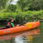 Boy in life vest paddles bright orange kayak on calm river surrounded by green trees.