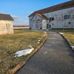 Old white barn and farmhouse on a sunny, rural landscape with a paved walkway.