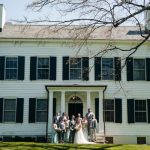 Wedding party stands in front of a classic white farmhouse under a bright sky.
