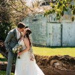Newlyweds kiss outdoors in a sunny, rustic setting with an old barn backdrop.