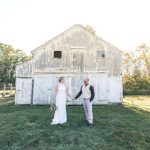 Bride and groom stand in front of a rustic white barn at sunset.