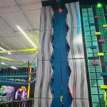 Indoor trampoline park with a large, wavy blue and white climbing wall feature.